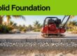 A red compactor presses crushed stone beneath aligned concrete pavers.