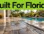 Textured patio pavers beside a turquoise pool with tropical plants and a visible drain.