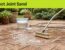 A brush and gentle stream of water clean brick pavers beside a bucket, leaving the sand-filled joints intact.