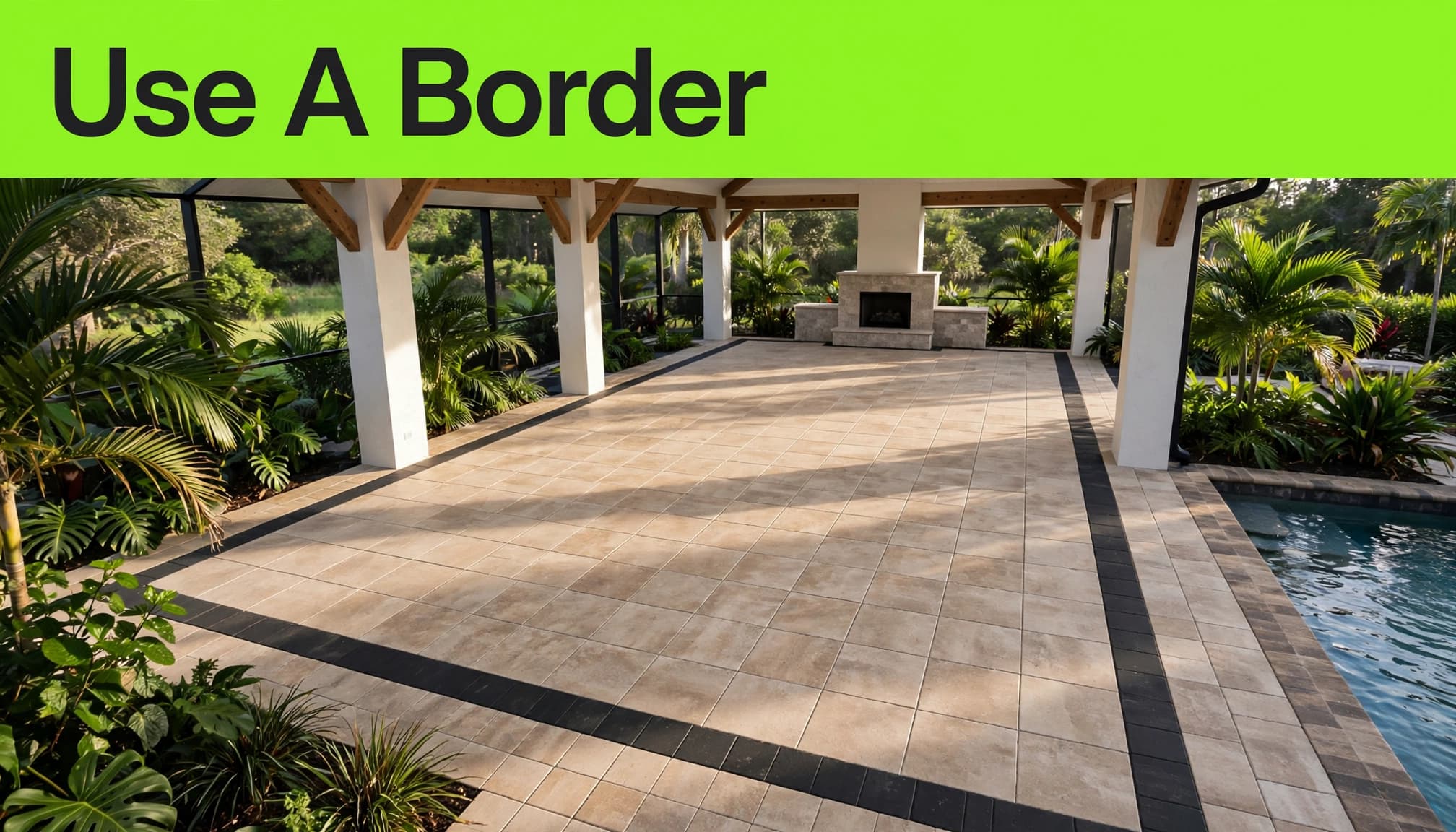 A stone patio with a contrasting paver border, fireplace, tropical plants, and pool edge.
